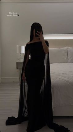 a woman in a black dress taking a selfie with her cell phone while standing next to a bed