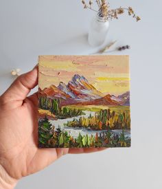 a hand holding up a small piece of art with mountains in the background and water below