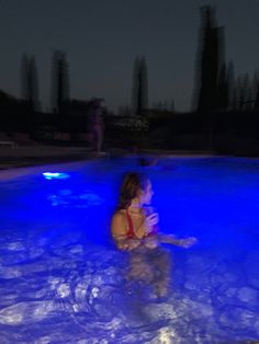 a woman sitting in the middle of a swimming pool at night with blue lights on it