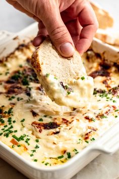 a hand dipping a piece of bread into a casserole dish with melted cheese and parsley