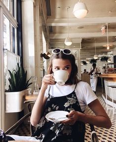 a woman sitting at a table drinking from a coffee cup and holding a plate in front of her face