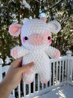 a hand holding a pink and white crocheted stuffed animal in front of snow covered trees