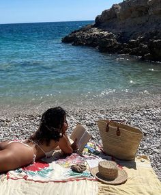 a woman laying on top of a beach next to the ocean reading a book and holding a straw hat