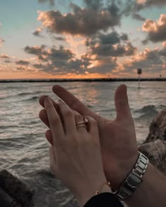 two hands holding each other near the water at sunset or sunrise with clouds in the background