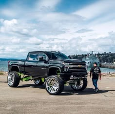 a man standing next to a black truck on top of a parking lot near the ocean