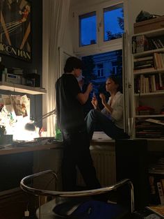 two people standing in a living room next to a book shelf with books on it