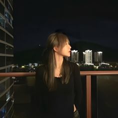 a woman standing on top of a balcony next to a tall building at night with city lights in the background