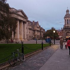 people walking down the sidewalk in front of some old buildings at dusk with lights on