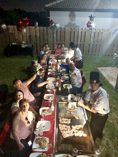 a group of people sitting at a long table with food on it in the backyard