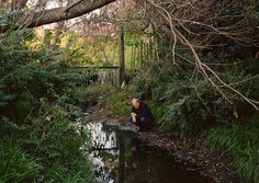 a man squatting down next to a river in the woods with trees and bushes around him