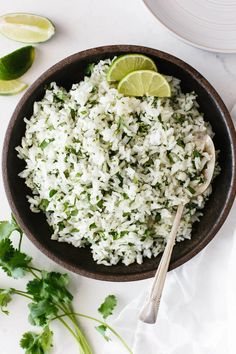 rice and cilantro in a bowl with limes next to it on a white surface