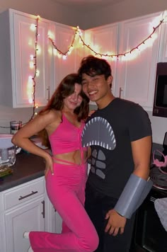 a man and woman standing in a kitchen with lights on the wall behind them, posing for a photo