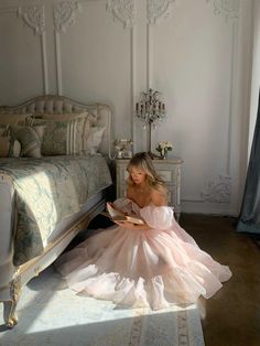 a woman in a pink dress sitting on the floor next to a bed and reading a book