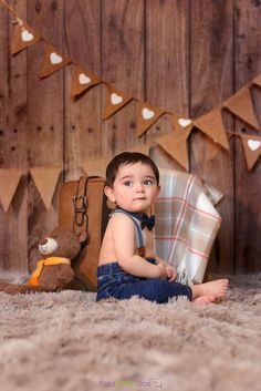 a little boy sitting on the floor in front of a wooden wall with bunting