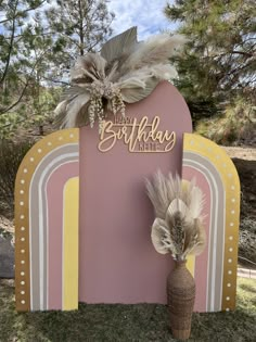 a pink and yellow birthday sign with feathers on it next to a vase filled with flowers