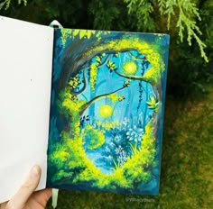 someone is holding up an art journal with trees and flowers painted on it in front of some green grass