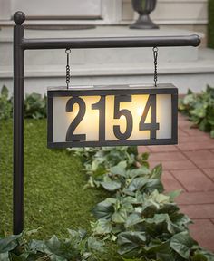 a house number sign hanging from the side of a building with grass and bushes around it