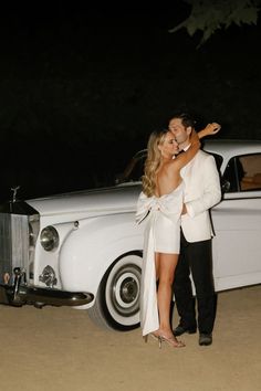 a man and woman standing in front of a white car at night with their arms around each other