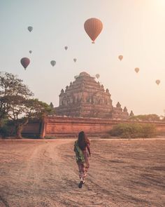 a woman walking down a dirt road with hot air balloons in the sky above her