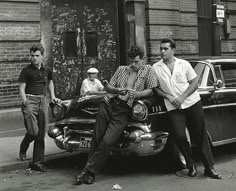 three men are sitting on the hood of an old car in front of a brick building