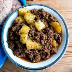 a bowl filled with meat and potatoes on top of a wooden table next to bread