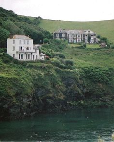 an old house on the side of a hill next to a body of water with ducks swimming in front of it