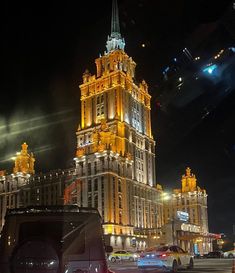 a very tall building lit up at night with cars parked in the street below it