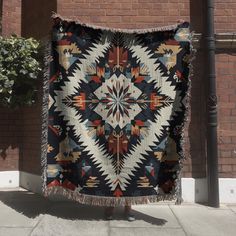 a person standing next to a brick building holding up a large quilt on the sidewalk