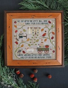 a cross - stitch sample in a wooden frame surrounded by pine cones and acorns