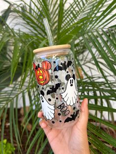 a hand holding up a cup with halloween decorations on it and a straw in front of a potted plant