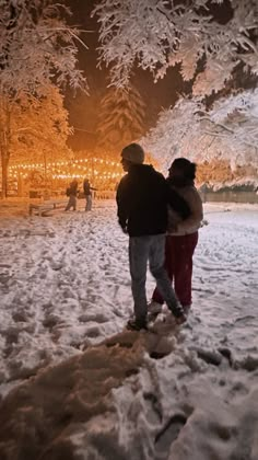 two people walking in the snow at night with lights on trees and buildings behind them