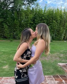 two women are kissing each other in front of some grass and trees, one is wearing a purple dress