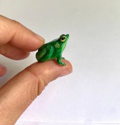 a small green frog figurine sitting on top of someone's finger in front of a white background