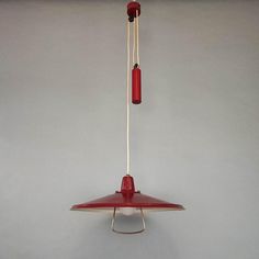 a red and white bird feeder hanging from a hook on a gray wall next to a light bulb