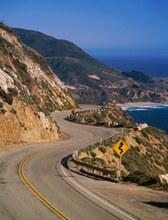 an empty road on the side of a mountain next to the ocean with a yellow sign