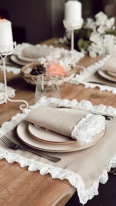 a wooden table topped with white plates and silverware covered in lace next to candles