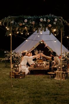 two people are sitting at a table in the middle of a field with lights strung over it