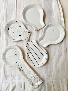 four white dishes with black speckles and spoons on a white cloth covered tablecloth