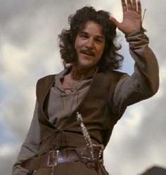 a man with long curly hair waves to the sky while standing in front of clouds