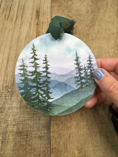 a hand holding a round ornament with trees on it and mountains in the background