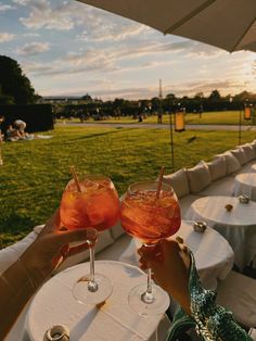 two people are holding up wine glasses at an outdoor table with white clothed tables and grass in the background