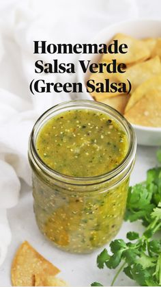 a glass jar filled with green salsa next to chips and cilantro on a white cloth