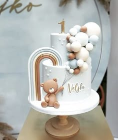 a teddy bear on top of a white cake with balloons and a rainbow in the background