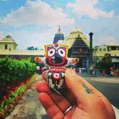 a hand holding a small toy in front of a building with many trees and bushes