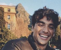 two men are smiling for the camera in front of a building with mountains behind them