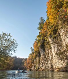 two people in a boat on a river surrounded by trees with fall foliage around them