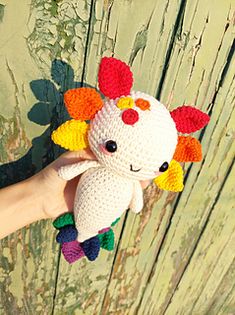 a hand holding a crocheted stuffed animal in front of a wooden fence with leaves on it