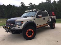 an off road truck with orange rims parked in a parking lot next to trees