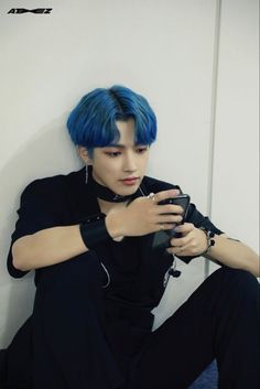 a young man with blue hair sitting on the floor looking at his cell phone while wearing black