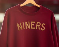 NINER FAITHFUL! Boost your sideline style with our classy San Francisco 49ers Crewneck Sweater, designed expressly for those who hold the gold and red close to their hearts. Essential gear for every Niners devotee, this sweater is more than just for those touchdown moments; it seamlessly elevates any relaxed ensemble, ensuring you're the star of every fan festivity. Whether you're channeling the grit of George Kittle or reminiscing legends like Joe Montana and Jerry Rice, our apparel has you outfitted in glory. From the iconic hills of San Francisco to spirited game day celebrations, GO NINERS! Every Sunday? Set the fashion gold standard! Note: Primarily curated for our 49ers' ladies, this sweater's unisex vibe is equally captivating on gents. Because Niners pride knows no bounds! 😉 👕🧢 49ers Outfit, 49ers Svg, Homemade Shirts, George Kittle, Nick Bosa, Brock Purdy, Sunday Football, Joe Montana, Football Sunday